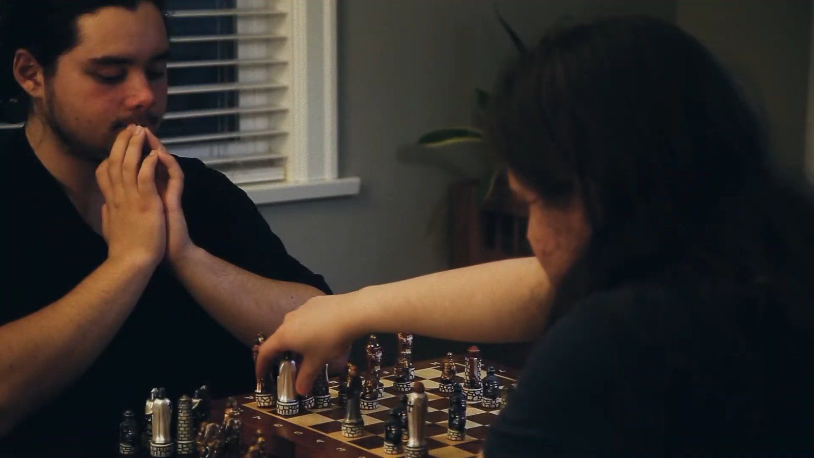 The two teens playing chess indoors, one reaching to make a move